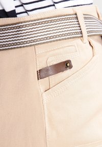 Los pantalones beige presentan un cinturón a rayas, un pequeño detalle de cuero marrón en el bolsillo y una tela suave y texturizada.