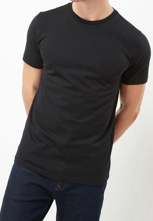 Homme portant un t-shirt noir uni à manches courtes et un jean bleu foncé, debout les mains dans le dos devant un fond blanc.