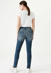 Vaqueros ajustados de denim azul medio, con bolsillos traseros, un ligero desvanecimiento y un diseño entallado. Combinado con una camiseta blanca básica y zapatillas.