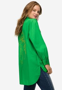 Camicia verde a maniche lunghe realizzata in tessuto morbido con una linea gialla disegnata sul retro. Orlo curvo e polsini con bottoni.