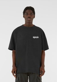 Tricou oversize negru din bumbac moale, cu guler rotund și mâneci scurte. Pe partea din față are un logo cu text alb pe zona din stânga a pieptului.