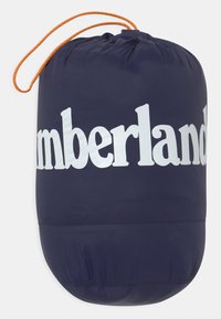 Marinblå dragkedjesäck med vitt "Timberland"-logotyp. Tillverkad av lätt material, har en texturerad yta och orange dragkedja.