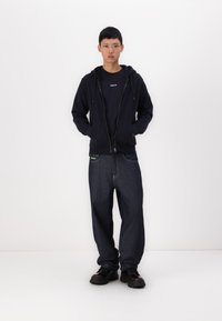 Mornarska zip-up kapuca z žepi in črnim napisom, v kombinaciji z ohlapnimi temnimi kavbojkami. Obutev so debele črne čevlje.