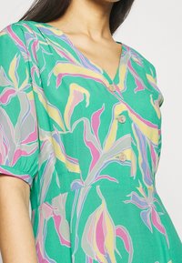 Robe verte avec un décolleté en V, des manches courtes bouffantes et une fermeture à boutons. Décorée de motifs floraux abstraits en rose, jaune et gris.