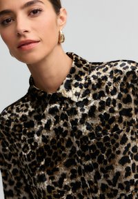 Leopardmønstret skjorte i blødt, glansfuld stof. Har krave, sorte knapper og en brystlomme, med en blanding af brune nuancer og sorte pletter.