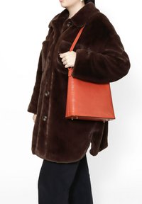 Cappotto in finto pelliccia marrone con grandi bottoni, indossato sopra pantaloni scuri. Una borsa rossa con texture è tenuta da una mano, caratterizzata da una forma triangolare e piedini dorati.