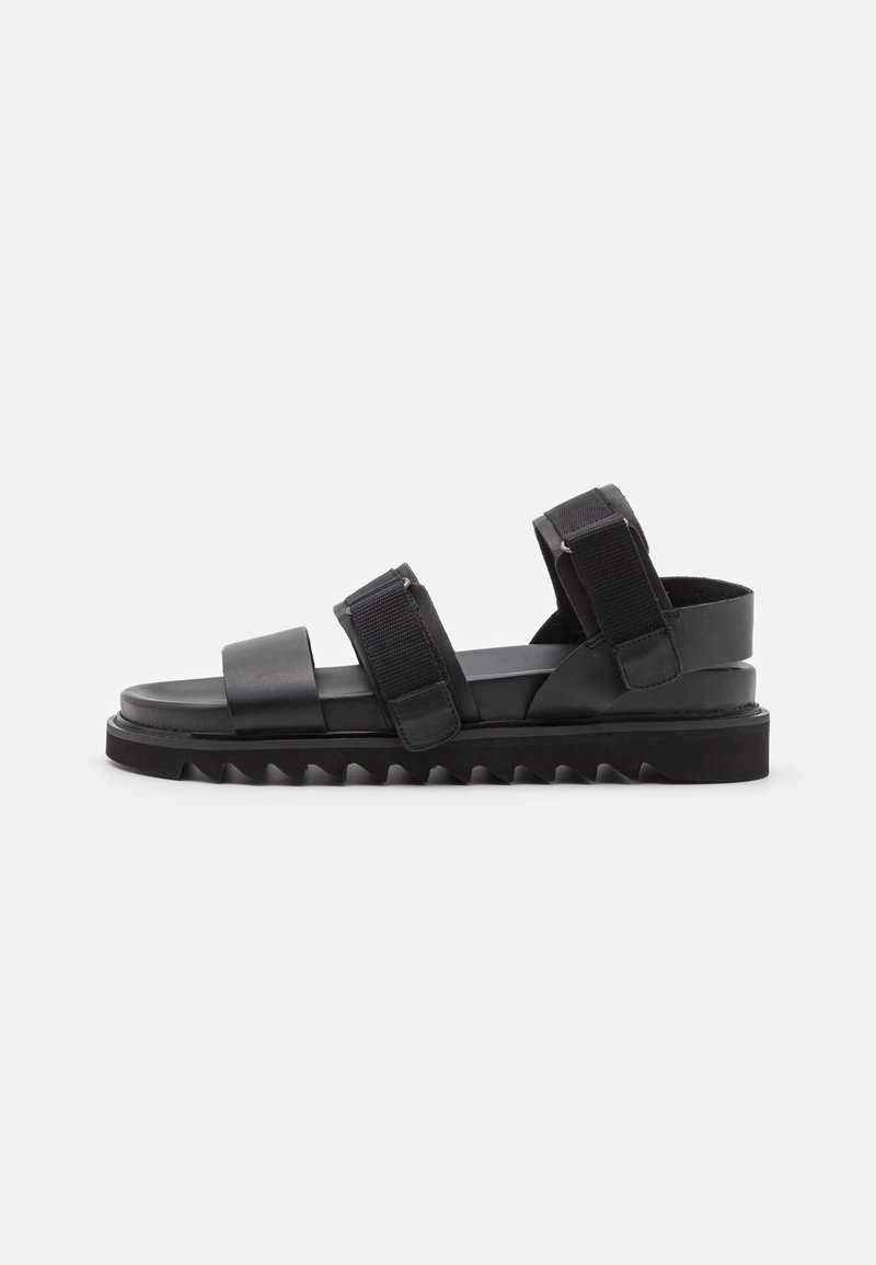 Sandal preto com duas tiras largas com fecho de velcro, sola plana e fundo texturizado em zigue-zague para melhor aderência. Acabamento em couro liso.