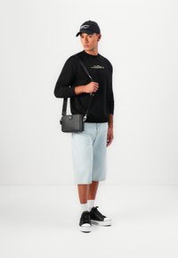 Sweatshirt preto com letras douradas, calções de ganga azuis claros, ténis pretos e uma pequena bolsa preta de ombro.
