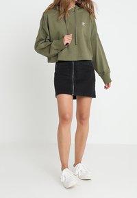 Olivgrön kort hoodie, svart denimkjol med framdragkedja och vita sneakers. Tyget verkar mjukt med en avslappnad design.