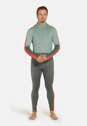 Mężczyzna stoi boso, ubrany w zielone i szare legginsy sportowe oraz pasującą długą bluzę z pomarańczowymi paskami, z rękami złożonymi przed sobą.