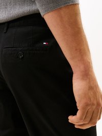 Calças pretas com um bolso traseiro com botão, apresentando um pequeno logo vermelho, branco e azul. O tecido parece suave com um acabamento mate.