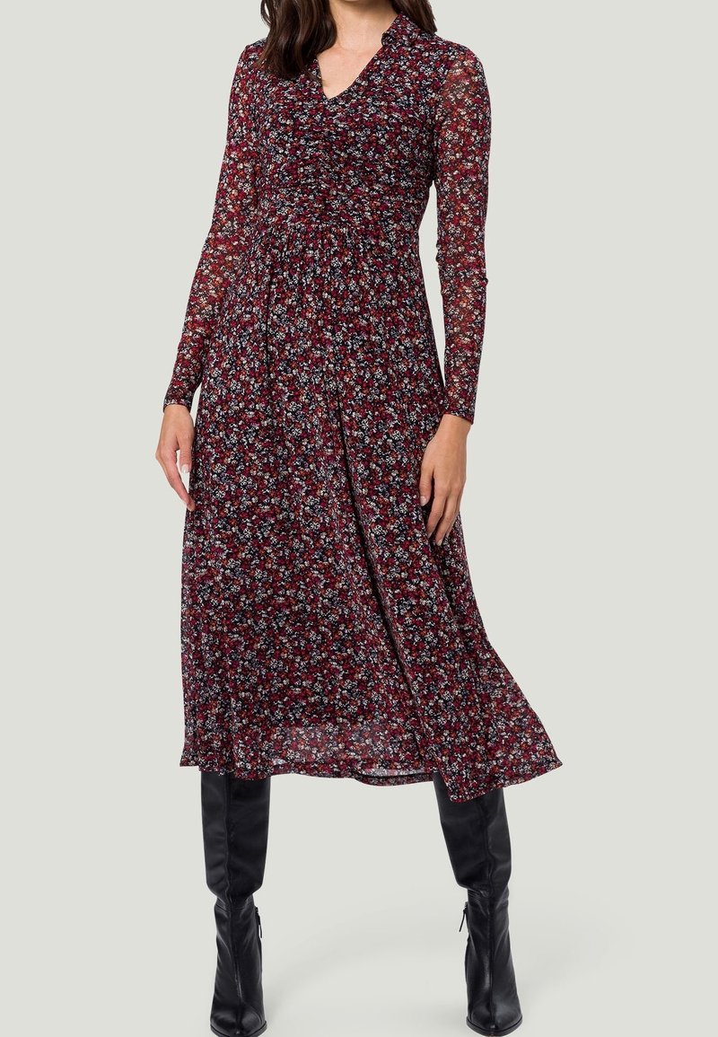 Långärmad blommig klänning med djup V-ringning, med en mörkröd bakgrund, små blå och vita blommor samt en svepande, veckad design.