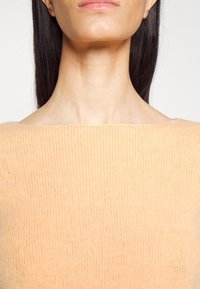 Närbild av en persons nacke och nedre ansikte som bär en mjuk, persikofärgad stickad tröja med off-the-shoulder halsringning.