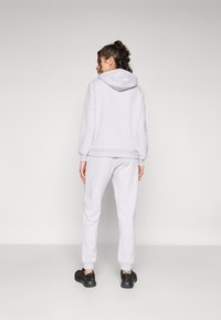 Svijetlo siva hoodie s kapuljačom na žnjor, rebrastim manžetama i odgovarajućim trenerkama. Teksturirani materijal, opušteni kroj i suptilni detalji brendiranja.