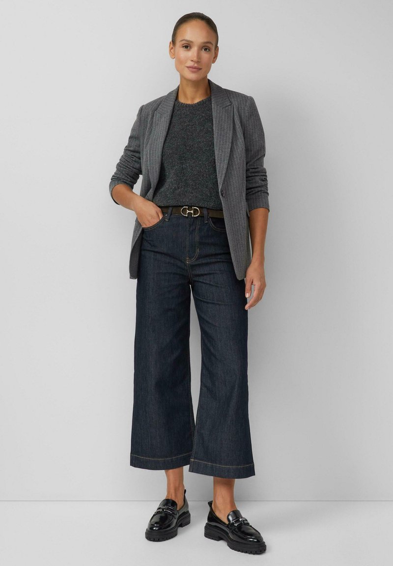 Blazer grigio a righe verticali su un maglione scuro, jeans denim scuri a gamba larga, mocassini neri e una cintura nera con fibbia d'argento.