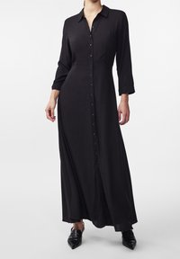 Abito maxi nero con bottoni a maniche lunghe, colletto e una texture liscia. Presenta una chiusura frontale con bottoni e fessure laterali.