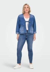 Denim jakna z v-izrezom, zavezanjem spredaj, dvema sprednjima žepoma in zavihanimi rokavi, v kombinaciji z denim hlačami in belimi supergami.
