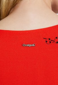 Czerwony element garderoby z zaokrąglonym dekoltem, gładką fakturą, srebrnym logo "Desigual" oraz czarnymi abstrakcyjnymi wzorami w okolicy ramienia.