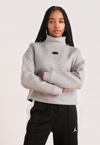 Felpa grigia corta con collo alla francese e piccolo logo nero, abbinata a pantaloni della tuta neri con strisce bianche sui lati.