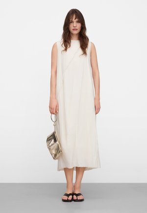 Frau mit langem braunem Haar, die ein weißes ärmelloses Midikleid, schwarze Sandalen trägt und eine metallisch goldene Handtasche mit rundem Griff hält.