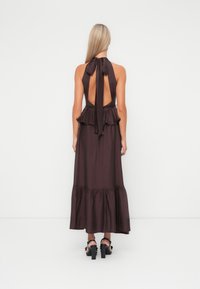 Ärmelloses braunes Maxikleid mit Halterneck, offenem Rücken, gerafftem Taillenbereich und ausgestelltem Saum. Accessoires mit schwarzen Riemchenschuhen.