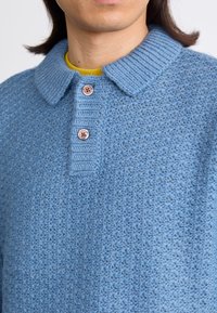 Μπλε πλεκτό πόλο πουλόβερ με ριγωτό γιακά, τριπλή κουμπότρυπα και υφή. Το ύφασμα φαίνεται να είναι μαλακό και ελαφρύ.