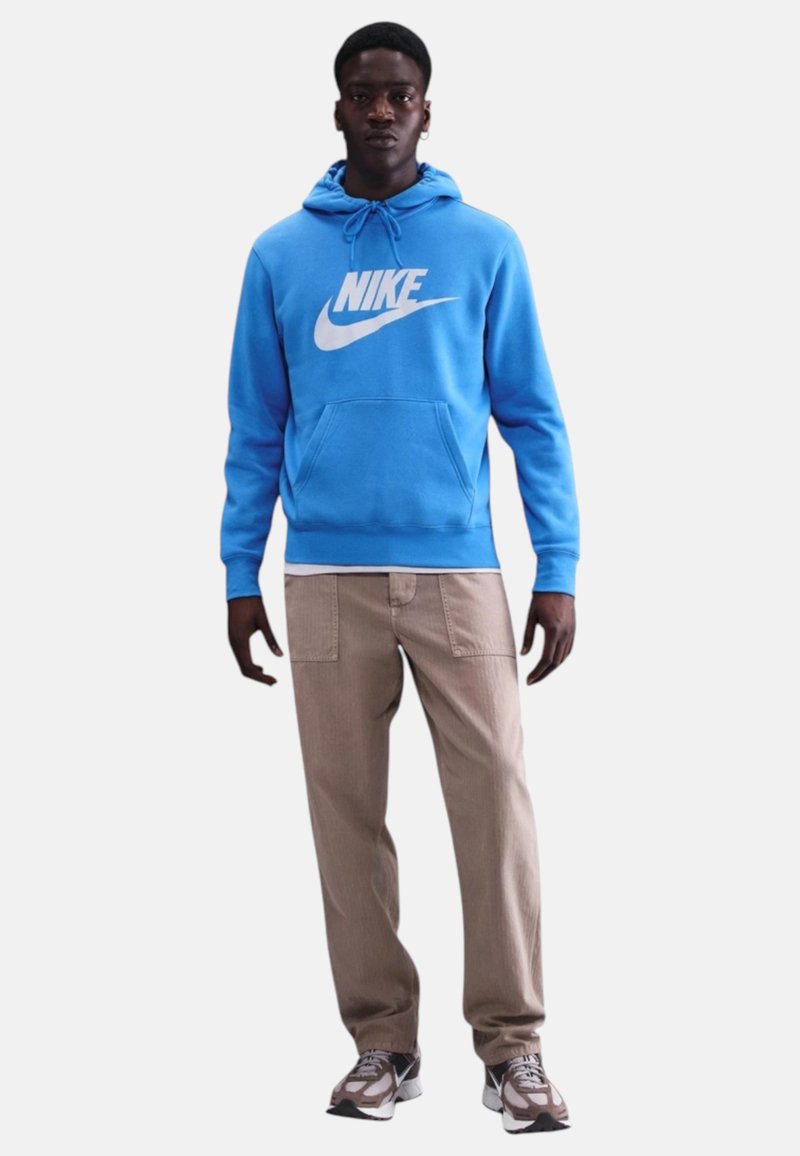 Uomo che indossa una felpa con cappuccio Nike blu brillante, pantaloni beige con tasche applicate e scarpe da ginnastica Nike grigie, in piedi contro uno sfondo bianco uniforme.