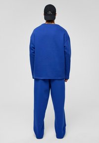 Blauwe sweatshirt en bijpassende broek van zacht materiaal, met een ontspannen pasvorm. De sweatshirt heeft een ronde hals. Zwarte pet weergegeven.