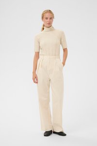 Kvinde med blond hår samlet bagpå, iført en beige turtleneck sweater med korte ærmer, højtaljede cremefarvede bukser og sorte sko, stående.