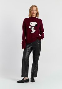 Bordeauxfarbener Strickpullover mit schwarzer Snoopy-Grafik; kombiniert mit schwarzen Lederhosen und schwarzen Slippern. Wolltextur, gerippter Kragen und Bündchen.