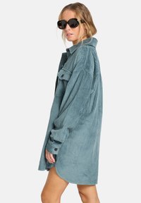 Lichtblauw corduroy jurk met lange mouwen, geknoopte manchetten, een kraag en een afgeronde zoom. Heeft een verticale ribbeltextuur.