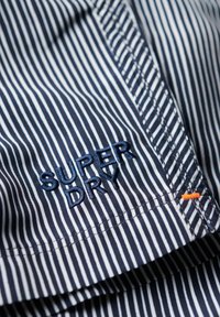Camisa de tela a rayas con una combinación de colores en azul marino y blanco. Logo bordado de "SUPER DRY" en azul oscuro con acento naranja. Textura suave.