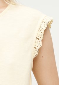 Κρεμ τοπ χωρίς μανίκια με λεπτή δαντέλα με floral μοτίβο στο άνοιγμα του χεριού, φορεμένο από άτομο με ανοιχτόχρωμο δέρμα και ξανθά μαλλιά.