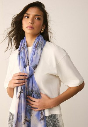 Young woman with long dark hair wearing gold hoop earrings, a white short-sleeve top, and a blue patterned scarf tied at the front.
