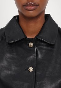 Veste en cuir noire brillante avec un large col et deux boutons dorés brillants, portée par une personne à la peau brune et aux lèvres brillantes.