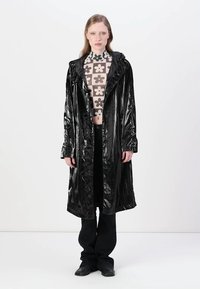 Černý, lesklý vinylový trenčkot s kapucí, předními knoflíky a bočními kapsami, kombinovaný s vzorovaným crop topem a černými kalhotami.