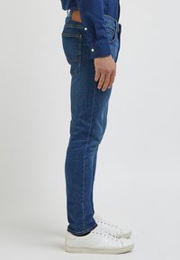 Siniset farkut kapealla istuvuudella, joissa on kaksi takataskua ja ruskea nahkapaikka vyötäröllä, yhdistettynä valkoisiin tennareihin.