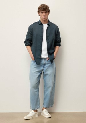 Tânăr în picioare cu mâinile în buzunare, purtând o cămașă închisă la culoare, nasturată, peste un tricou alb, blugi largi albaștri deschis și adidași albi.