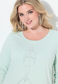 Uśmiechnięta kobieta o blond włosach nosi długie kolczyki z kryształkami oraz jasnomiętowy top z konturem modelek mody wykonanym z kryształków.