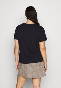 Mujer con cabello largo y oscuro que lleva una camiseta negra de manga corta y una falda de cuadros beige, de pie de espaldas a la cámara.