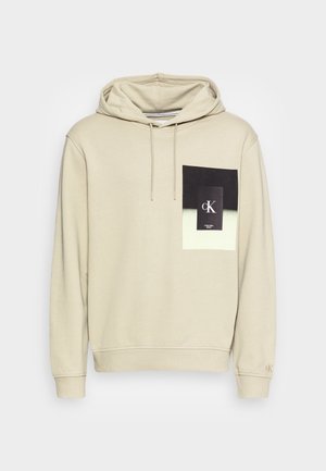 Beige hoodie met een zwart-witte vierkante afbeelding op de borst met de letters cK en de tekst Calvin Klein Jeans.