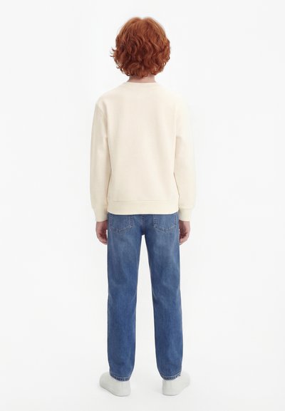 Persona con cabello rizado y rojo de espaldas, vistiendo una camisa de manga larga color crema, jeans azules y zapatos blancos contra un fondo blanco.