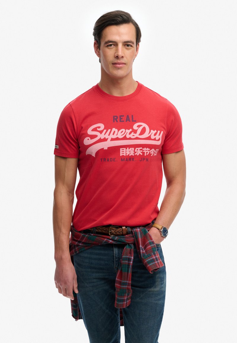 Junger Mann trägt ein rotes „SuperDry“-T-Shirt, blaue Jeans, ein kariertes Hemd um die Taille gebunden und eine Armbanduhr, steht vor weißem Hintergrund.