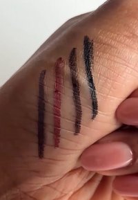 Quatre échantillons de maquillage sur une main : gris foncé, marron foncé, rouge profond et prune, montrant différentes épaisseurs et textures sur la peau.