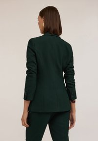 Blazer tailored di un verde scuro con spalle strutturate, maniche lunghe e bottoni decorativi sui polsini. Texture liscia e design aderente.