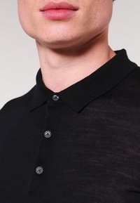 Nära bild av en persons nedre ansikte och nacke som bär en svart knäppt polo med klassisk krage.