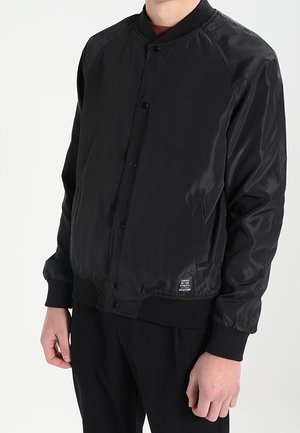 Mężczyzna ubrany w czarną kurtkę bomberkę z zatrzaskami, ściągaczami na mankietach i małą metką przy dolnym brzegu po lewej stronie.