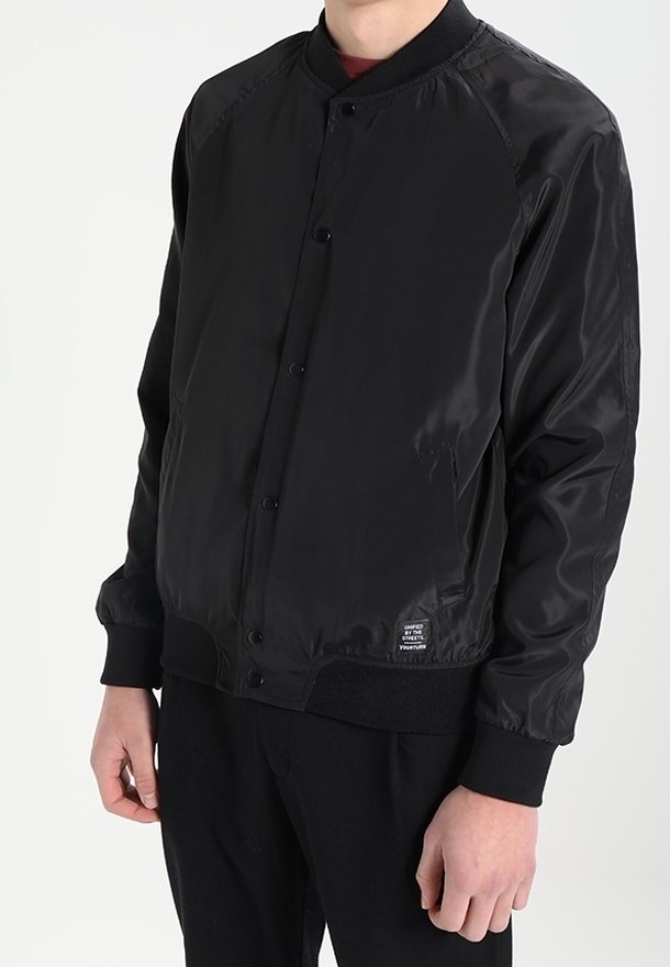 Homme portant une veste bomber noire avec des boutons pression, des poignets côtelés et une petite étiquette près de l'ourlet sur le côté gauche.
