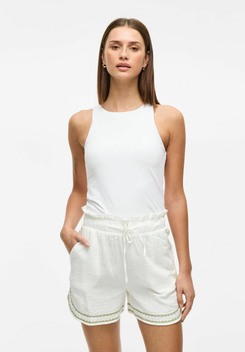 Haut sans manches blanc avec un col montant associé à un short blanc clair. Le short présente une taille élastique avec cordon et un liseré vert le long des ourlets.