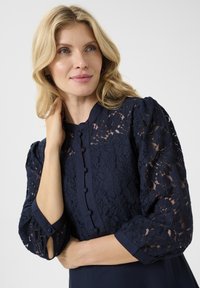 Navy blue dress featuring puffed, lace sleeves, button accents along the front, and a smooth fabric body. Textured lace detail provides contrast.